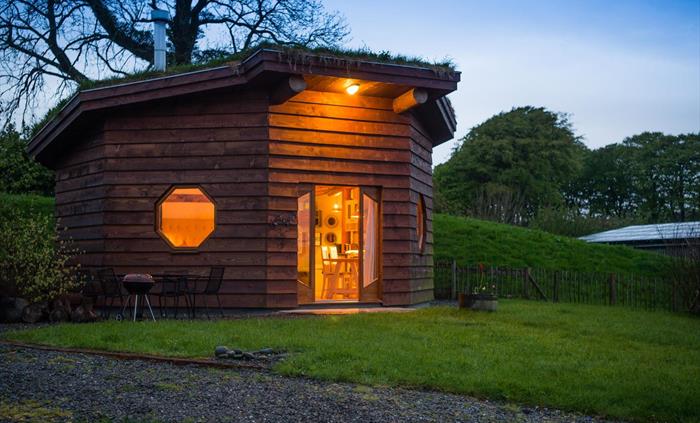 Saffir Lodge sits hidden in a wildflower meadow, its turf roof blending into the hills. Timber walls and warm windows glow beside a steaming wood-fire